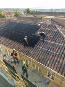 Operarios instalan paneles solares sobre un tejado de tejas, con arneses de seguridad y una plataforma elevadora.
