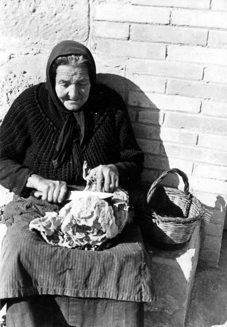 Coscojuela de Fantova saca su memoria a las calles con una exposición fotográfica Mujer mayor sentada cortando una hortaliza en una fotografía histórica de la exposición Lo silenzio que conta.