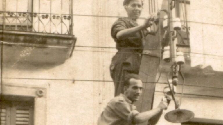 otografía antigua de dos operarios sobre una escalera trabajando en un tendido eléctrico junto a una fachada con balcones.