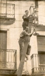 otografía antigua de dos operarios sobre una escalera trabajando en un tendido eléctrico junto a una fachada con balcones.